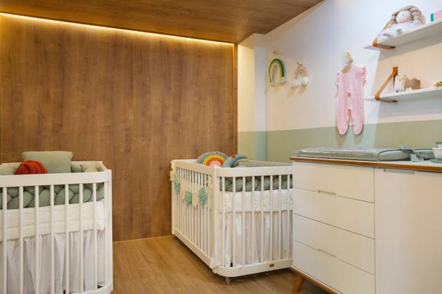 Um quarto de criança, com uma parede verde e outra em madeira, com dois berços brancos e um móvel de gavetas