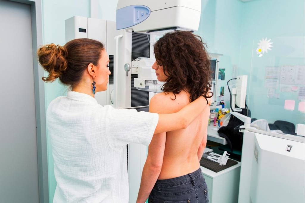 Uma mulher de cabelos pretos cacheados está sem blusa, de frente para uma máquina de mamografia, ao lado de uma médica usando jaleco branco