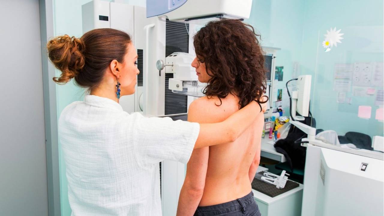 Uma mulher de cabelos pretos cacheados está sem blusa, de frente para uma máquina de mamografia, ao lado de uma médica usando jaleco branco