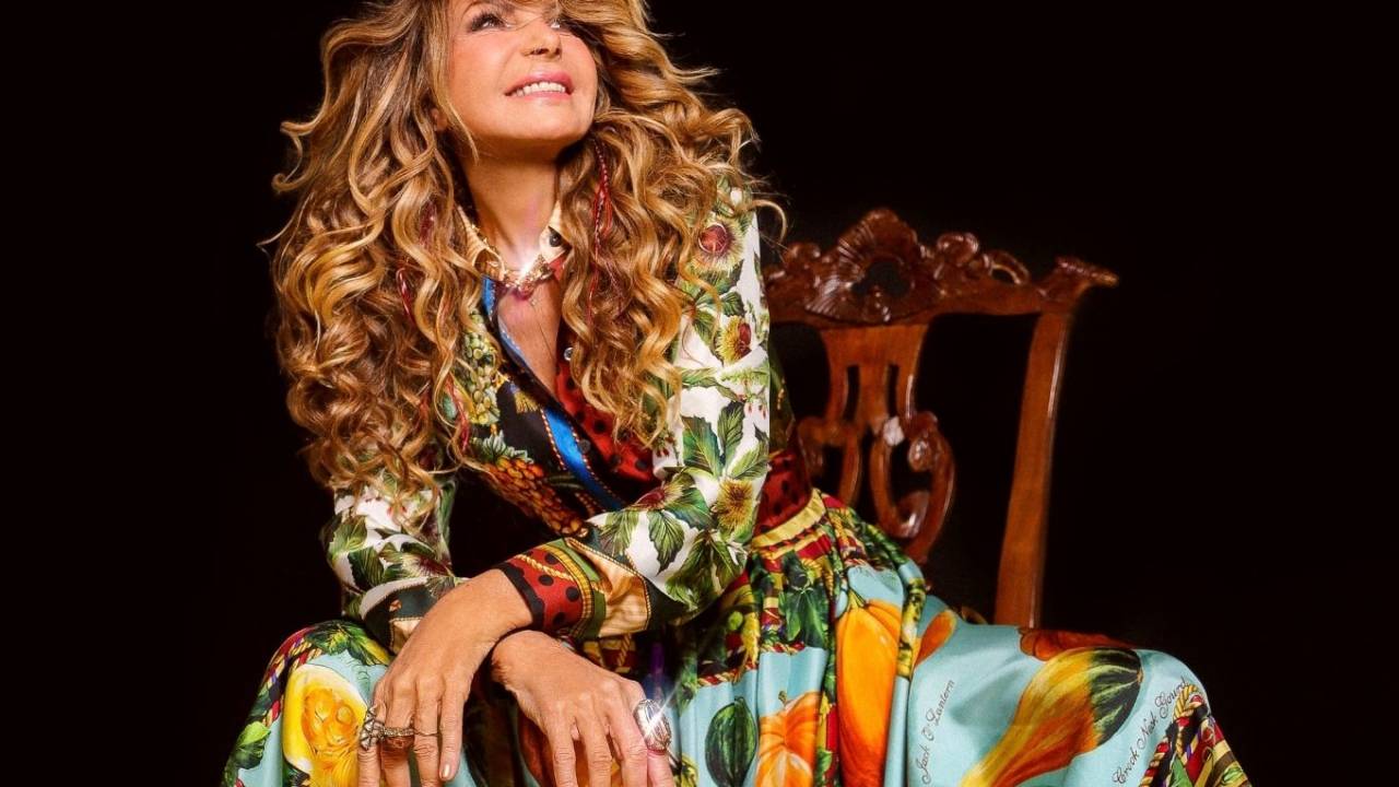 A cantora Elba Ramalho está sentada em uma cadeira olhando para o alto com um vestido todo estampado de magas compridas.