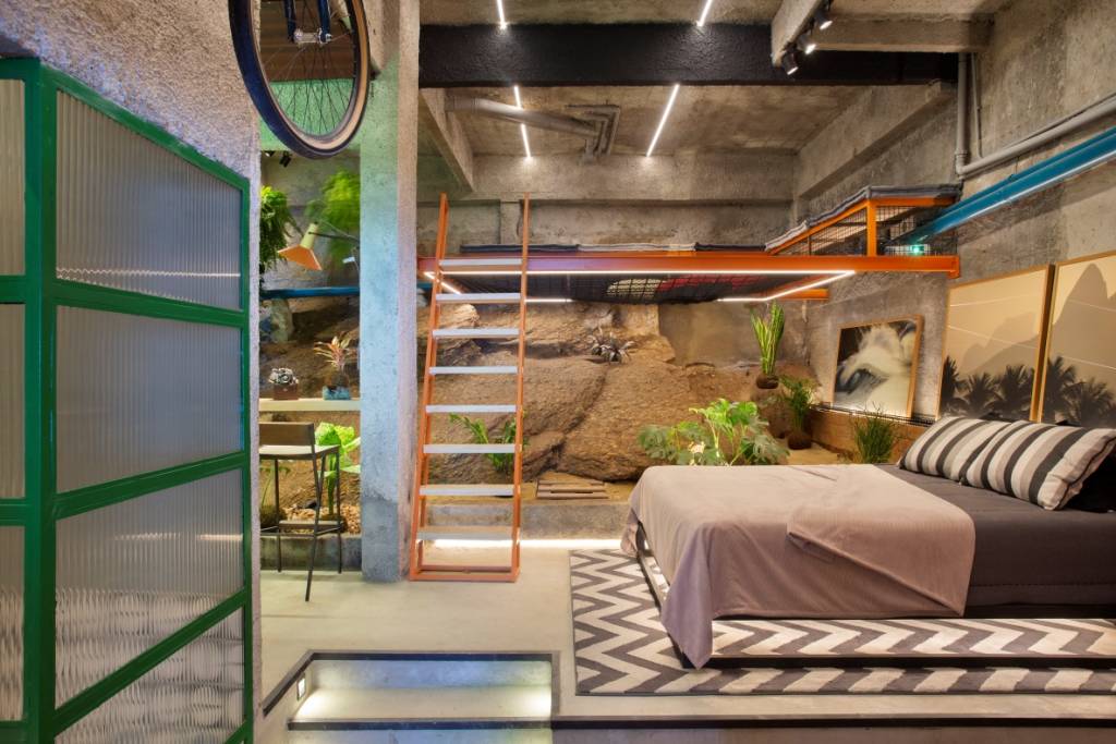 Um studio tem como uma das paredes uma grande pedra natural. Uma cama com lençol zinza está à direita e uma escada laranja leva para um mezanino à esquerda.