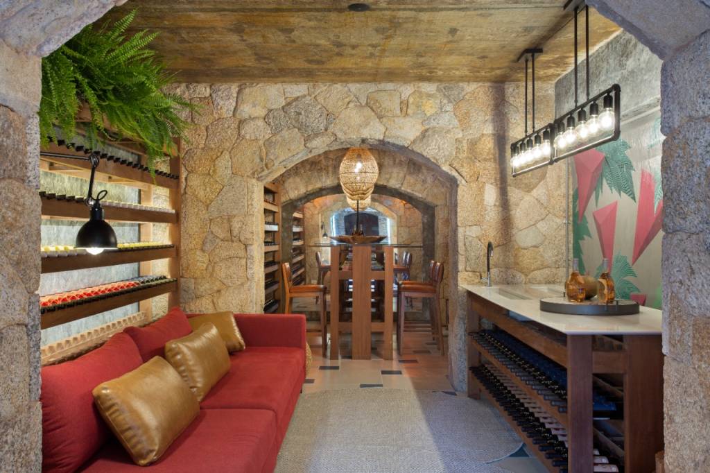 Uma dega de vinho em pedra tem um sofá vermelho à esquerda com almofadas de couro e uma bancada branca à direita