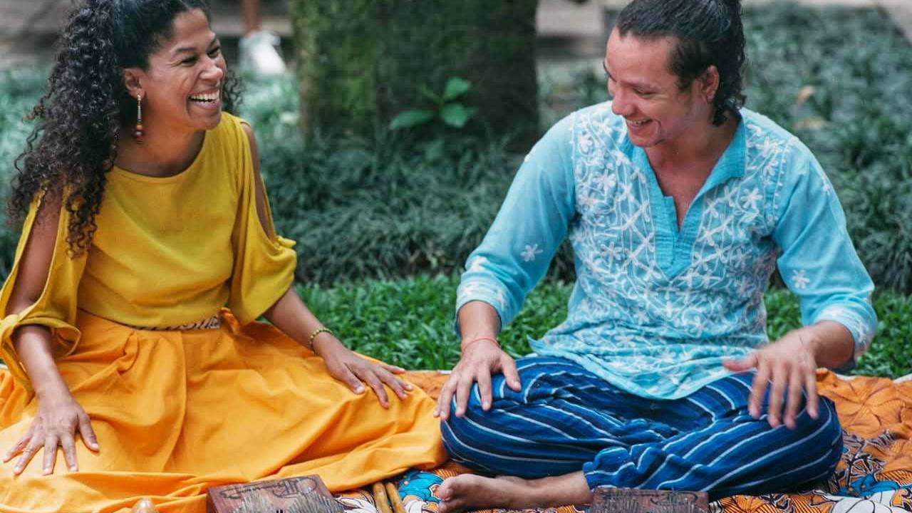 À esquerda, uma mulher negra de cabelos pretos cacheados com blusa e saia amarelas, sorrindo. Ao seu lado, também sorrindo, um homem branco com cabelo ondulado castanho, preso em um coque. sentado, vestindo bata azul-clara e calça com listras verticais azul-escuras e pretas. Os dois estão sentados sobre uma canga de fundo amarelo com flores azuis e brancas e detalhes vermelhos. À frente dele, instrumentos: um xequerê, duas kalimba e duas baquetas.