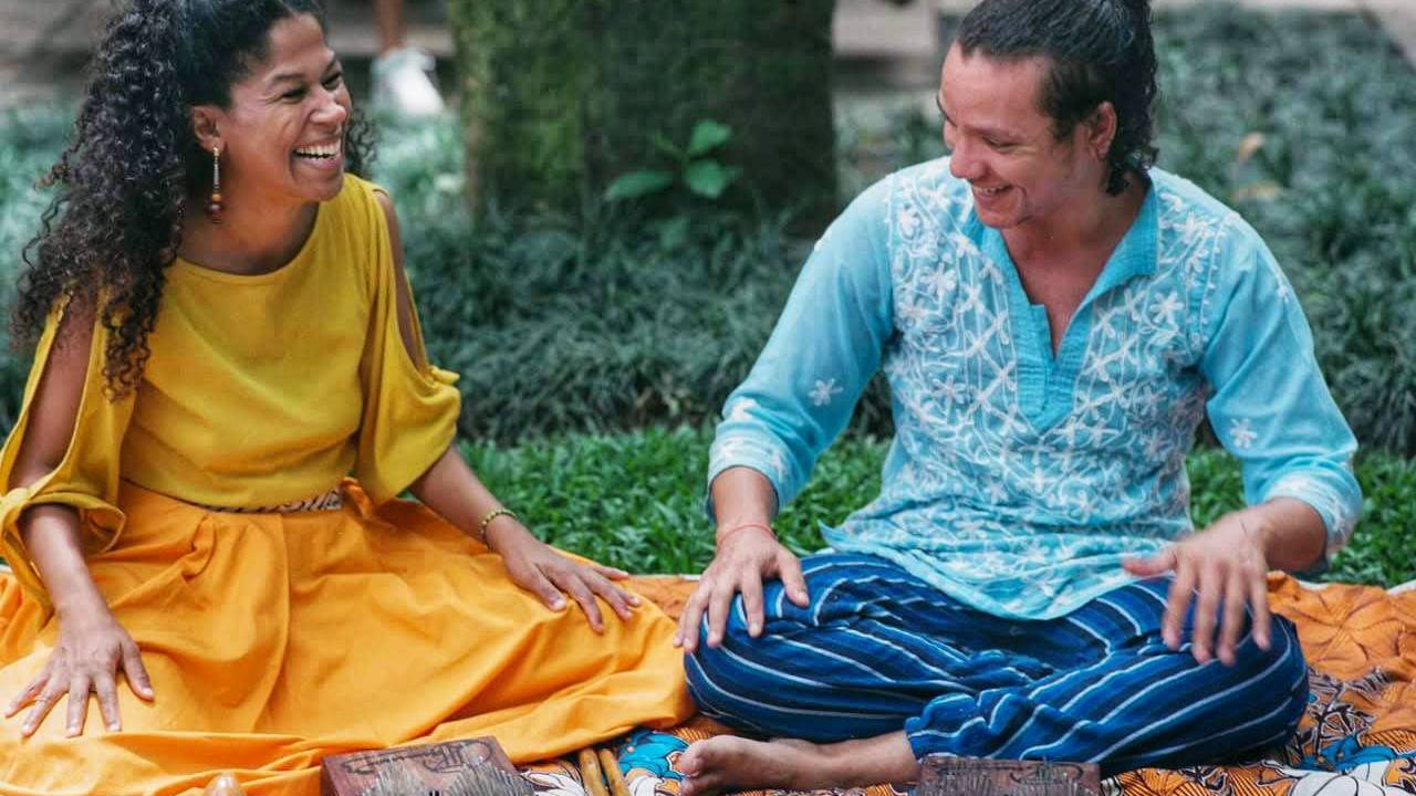 À esquerda, uma mulher negra de cabelos pretos cacheados com blusa e saia amarelas, sorrindo. Ao seu lado, também sorrindo, um homem branco com cabelo ondulado castanho, preso em um coque. sentado, vestindo bata azul-clara e calça com listras verticais azul-escuras e pretas. Os dois estão sentados sobre uma canga de fundo amarelo com flores azuis e brancas e detalhes vermelhos. À frente dele, instrumentos: um xequerê, duas kalimba e duas baquetas. 