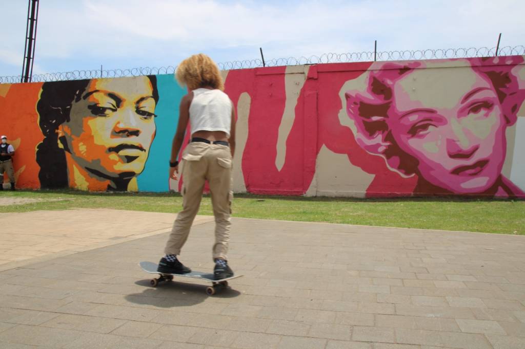 Foto mostra menina andando de skate em frente à mural pintado com mulheres famosas na história