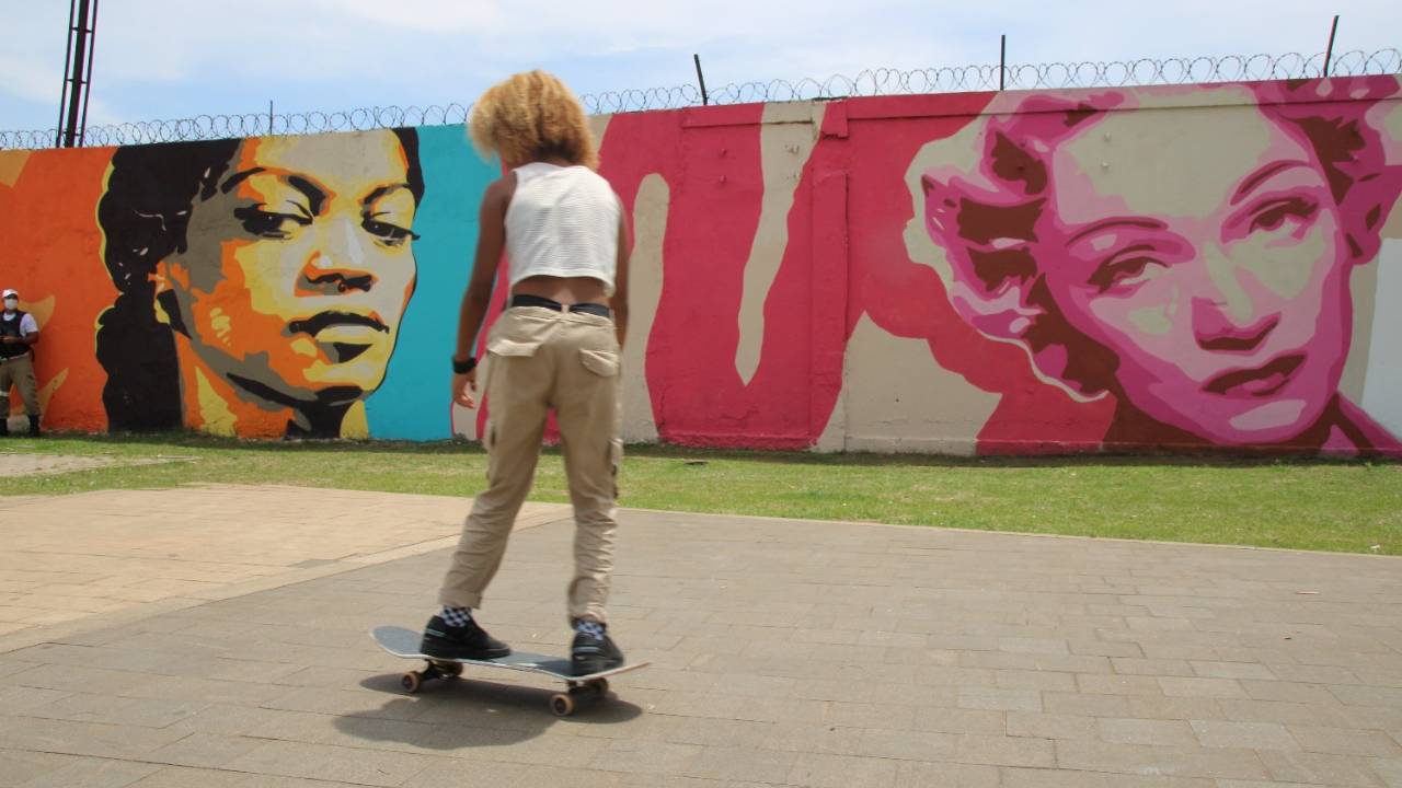 Foto mostra menina andando de skate em frente à mural pintado com mulheres famosas na história