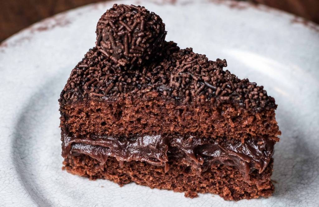imagem mostra bolo de chocolate recheado com brigadeiro e cobertura de chocolate com granulada e um brigadeiro enrolado em cima