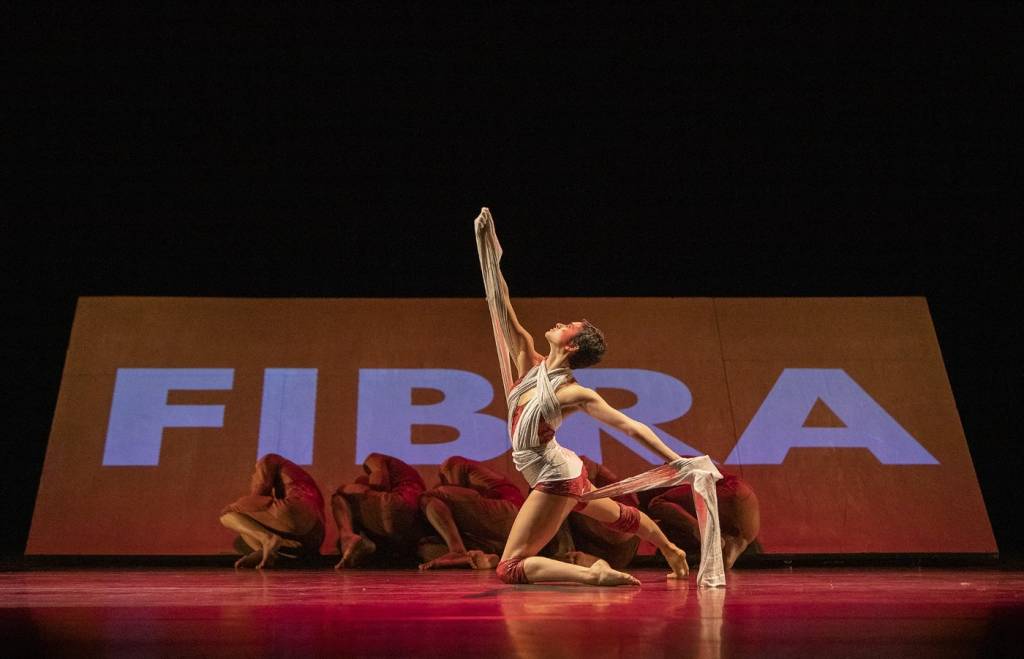 Bailarina da Cia Deborah Colker está no chão em primeiro plano. Em segundo plano, outros cinco bailarinos. Ao fundo, está projetada a palavra Fibra.