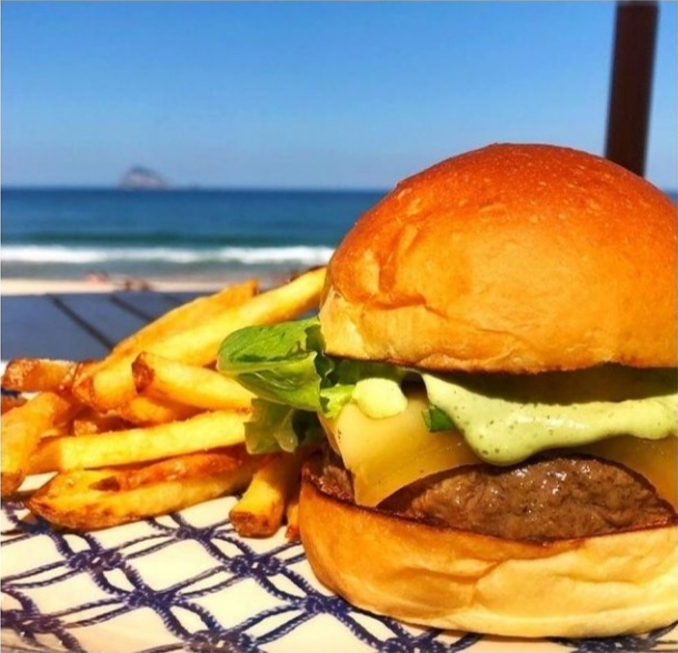 Foto mostra hambúrguer servido com batata frita com a praia ao fundo da imagem