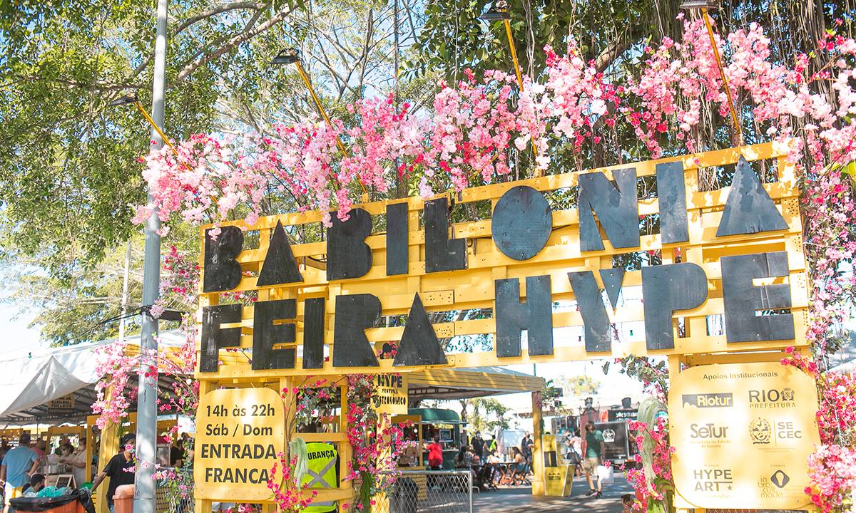 O pórtico de entrada da Babilônia Feira Hype, com letreiro preto sobre estrutura amarela e flores em cima do pórtico