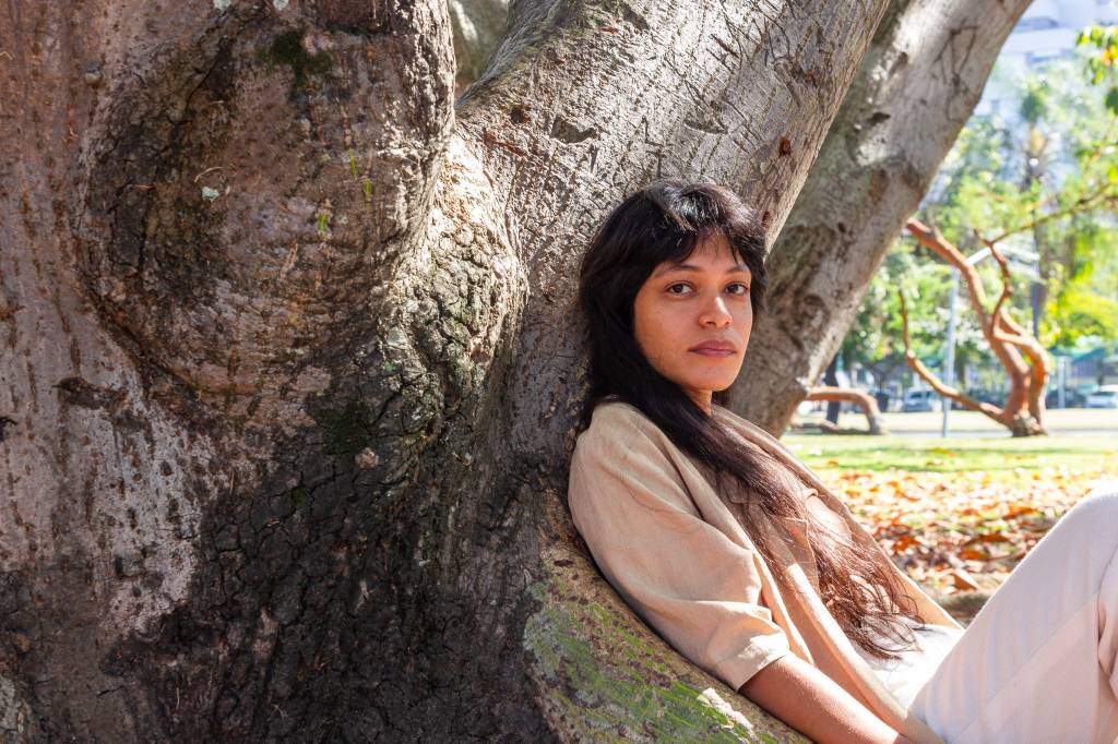 A artista plástica Sallisa Rosa usa roupa bege e está encostada em um tronco de árvore.