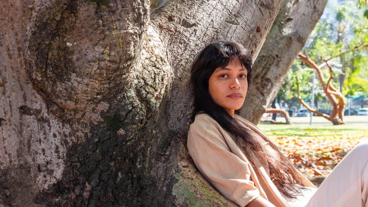 A artista plástica Sallisa Rosa usa roupa bege e está encostada em um tronco de árvore.