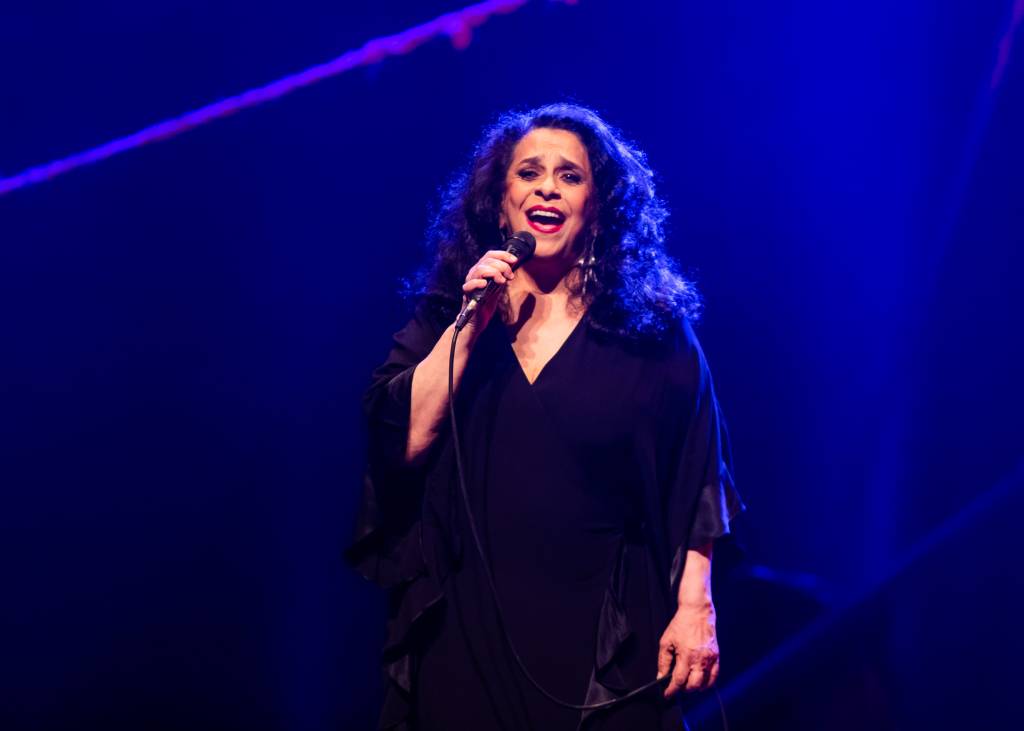A cantora Gal Costa cantando no palco, usando vestido preto. Ao fundo, uma luz azul