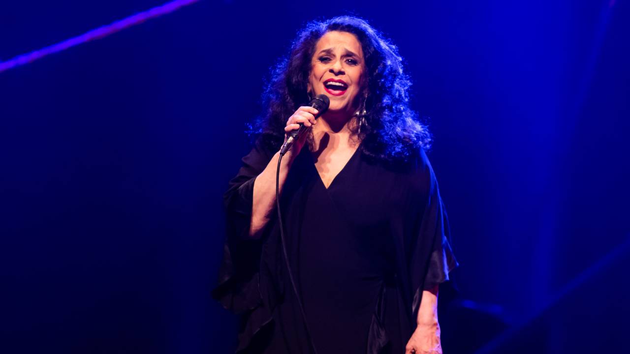 A cantora Gal Costa cantando no palco, usando vestido preto. Ao fundo, uma luz azul