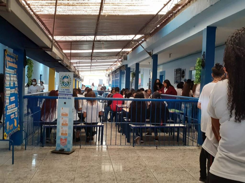 Foto mostra corredor de uma escola com as cores azuis com vários estudantes e um totem de alcool em gél