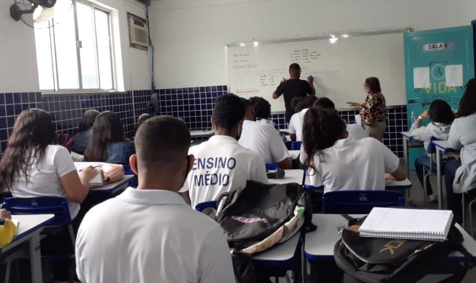 Foto mostra estudantes do ensino médio com uniforme branco em uma sala de aula com um professor à frente de um quadro branco