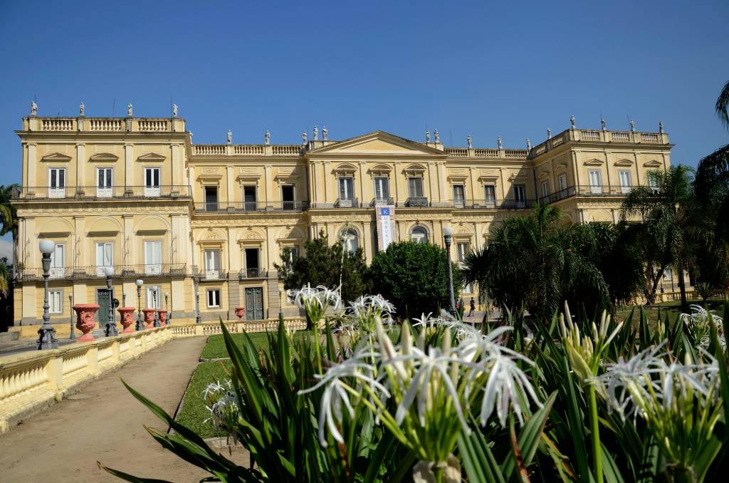 Atrás, a fachada do museu nacional, amarelada. Na frente, detalhe do jardim.