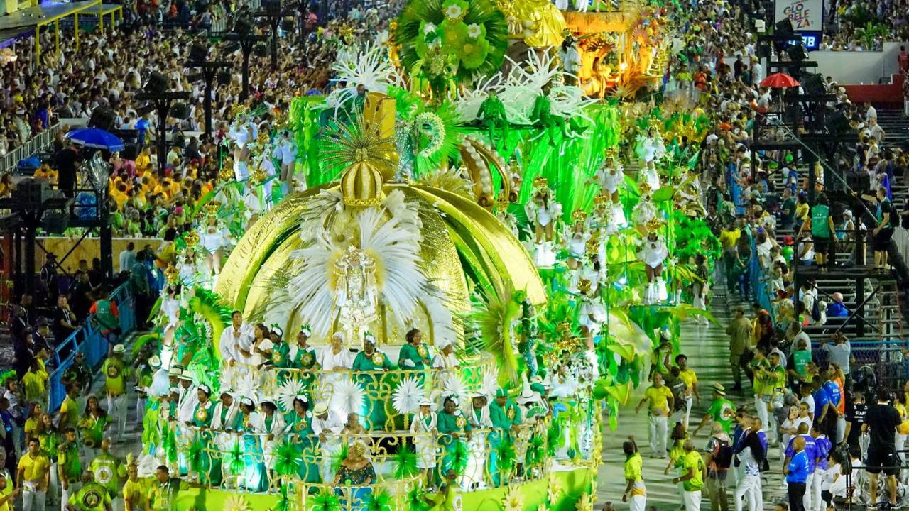 Carro abre-alas, com uma coroa no alto, e alas do Império Serrano, com predominância das cores verde e branca, fotografados de cima. Dos dois lados, a frente dos camarotes da Marquês de Sapucaí, lotadas.