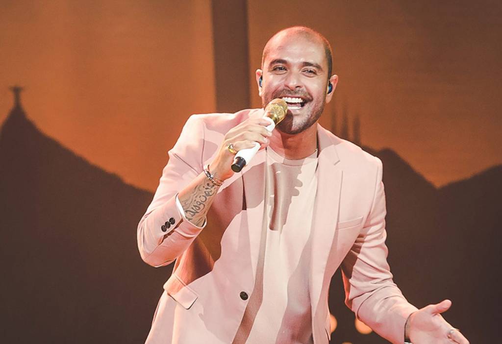 O cantor Diogo Nogueira cantando, vestindo terno e camisa rosa claros