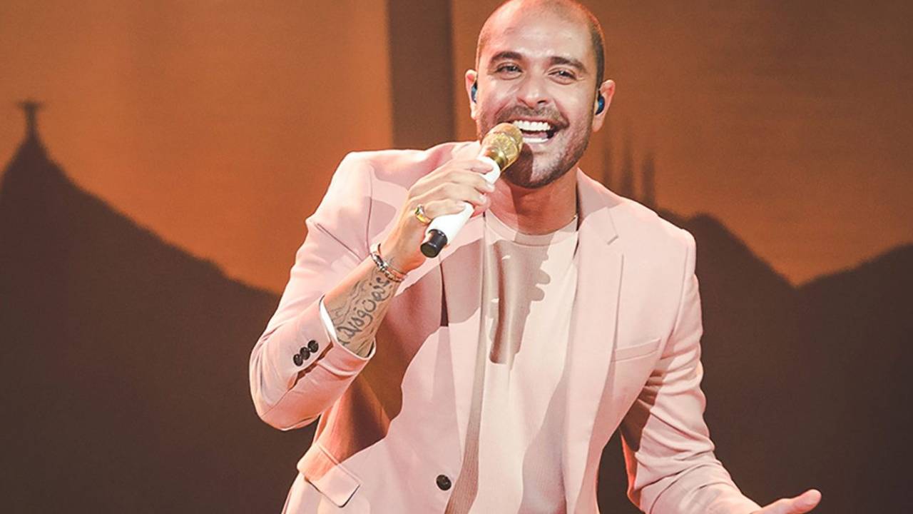 O cantor Diogo Nogueira cantando, vestindo terno e camisa rosa claros