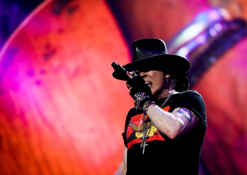 O vocalista do Guns N' Roses, Axl Rose, está cantando e usa chapéu preto e blusa preta com estampa vermelha e amarela
