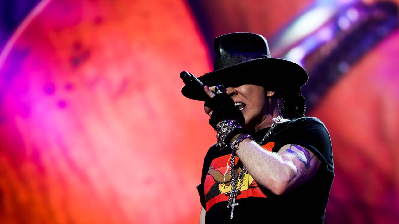 O vocalista do Guns N' Roses, Axl Rose, está cantando e usa chapéu preto e blusa preta com estampa vermelha e amarela