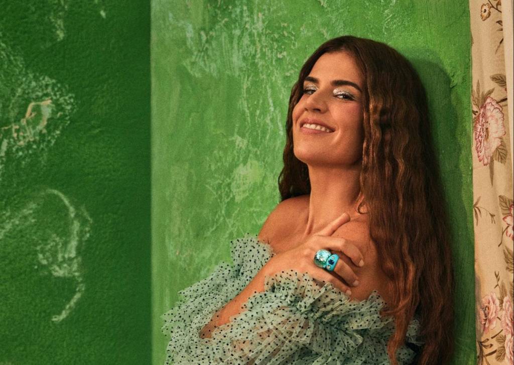A cantora Mariana Aydar está de lado, encostada em uma parede verde, sorrindo e olhando para a câmera. Ela usa vestido verde também.