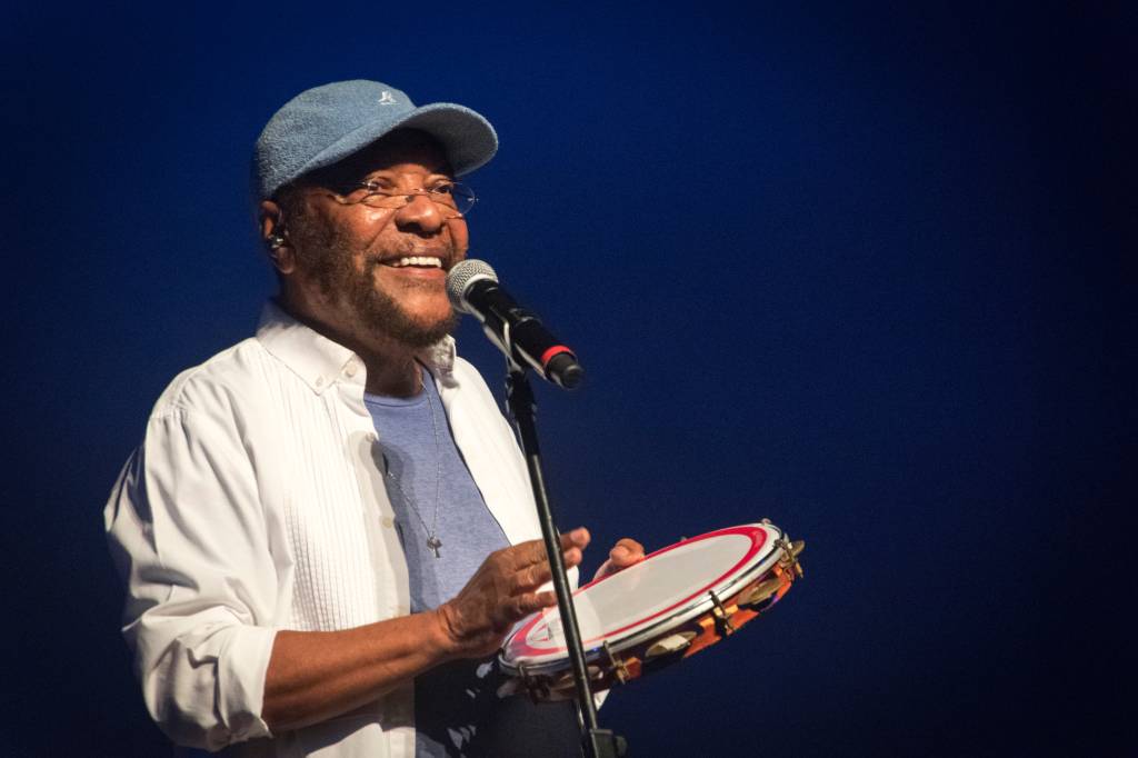 Martinho da Vila toca pandeiro no palco. Ele está de frente para o microfone. e veste blusa azul. camisa de manga comprida branca e boné azul.