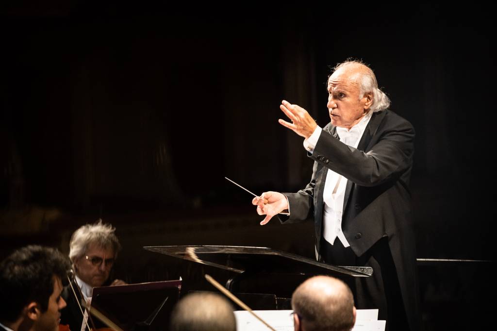 O maestro Isaac Karabtchevsky está à direita da foto, vestindo casaca preta e camisa branca, regendo a orquestra. Na foto, um está voltado para a câmera, o outro está de lado e os outros dois estão de costas.
