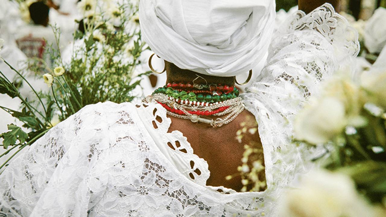 Fotografia de mulher praticante do candomblé com roupas brancas, de costas, usando turbante branco, com flores brancas na mão