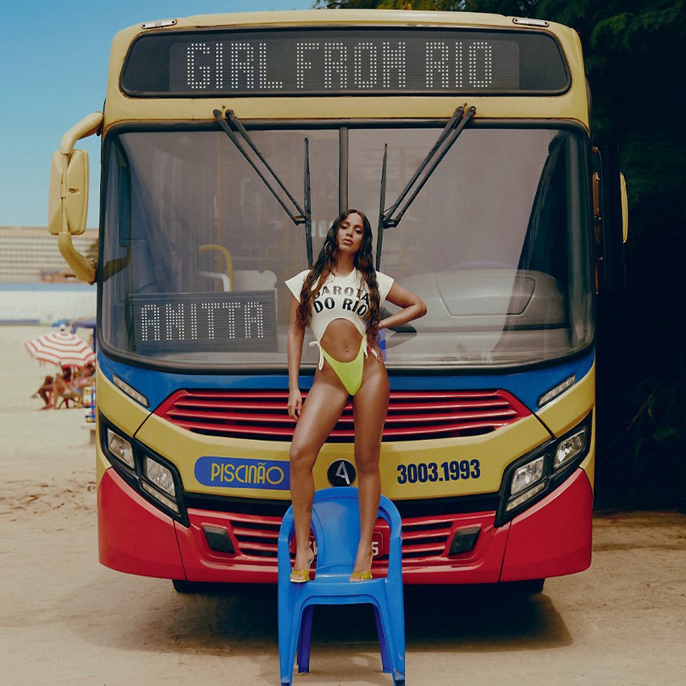 Anitta está em cima de uma cadeira azul e em frente a um ônibus com o letreiro escrito Girl from Rio