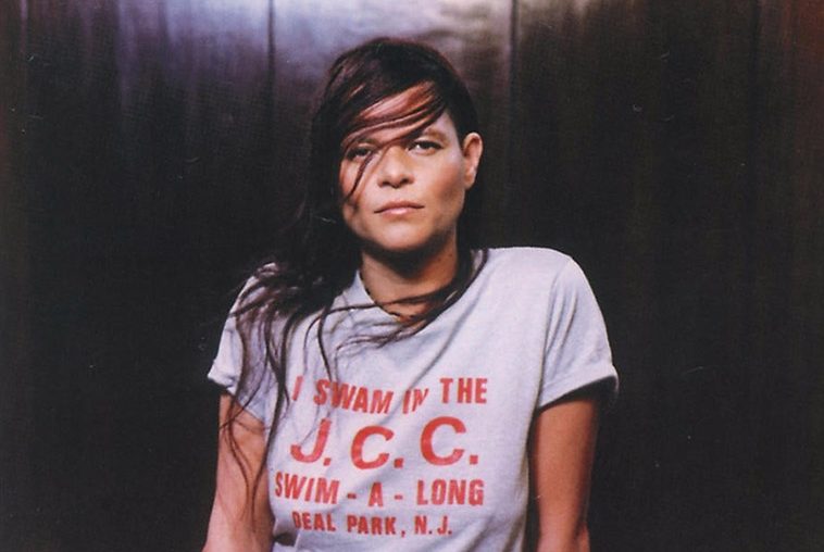 A cantora Cássia Eller com cabelo comprido, camiseta cinza com dizeres em vermelho. Ao fundo, uma parede de madeira.