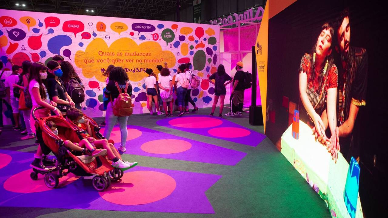 Foto mostra um espaço colorido, com um projeção de um vídeo e uma mãe com um carrinho de bebê e uma criança observando