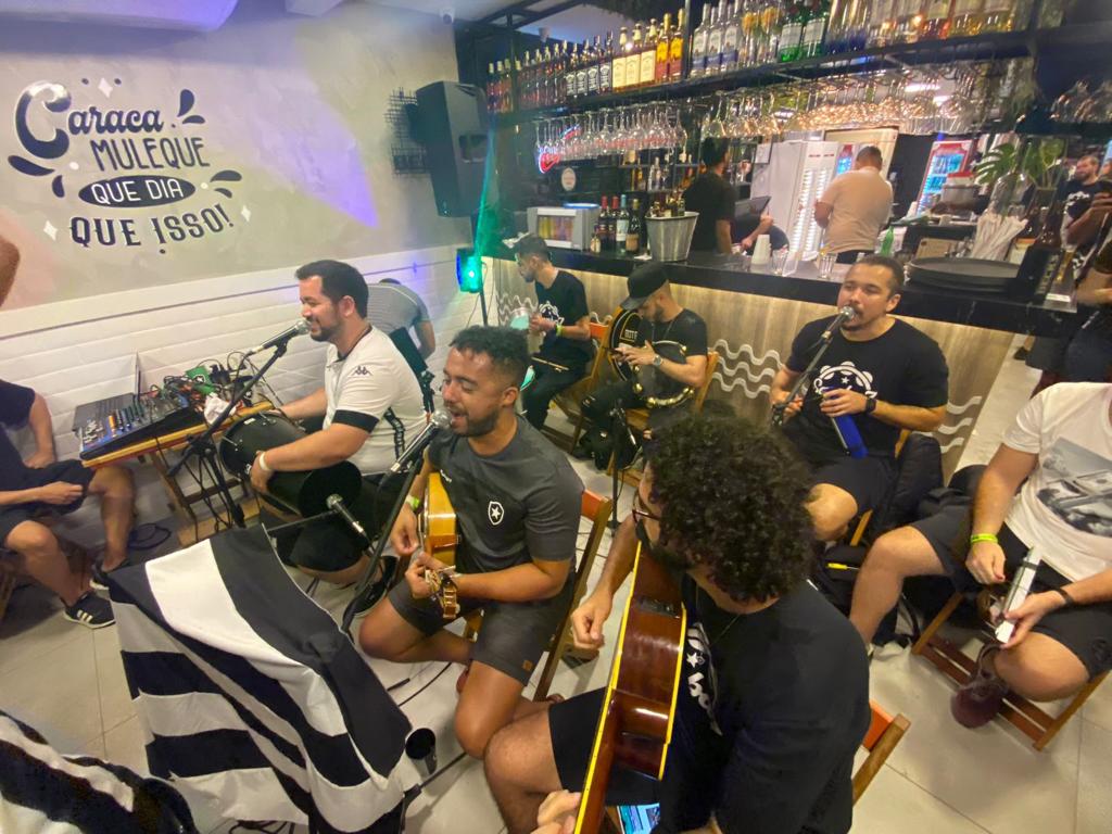 Grupo de música se apresenta dentro de um bar. Os sete integrantes, três na frente e quatro atrás, estão sentados e com microfones à frente. O piso do bar é preto e banco.