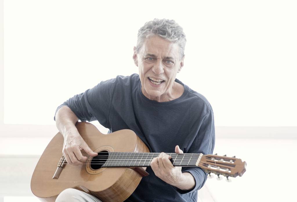 O cantor e compositor Chico Buarque está sentado em pose de quem toca o violão e com a boca entreaberta. Ele veste blusa azul-escuro e apoia o violão em um dos joelhos.