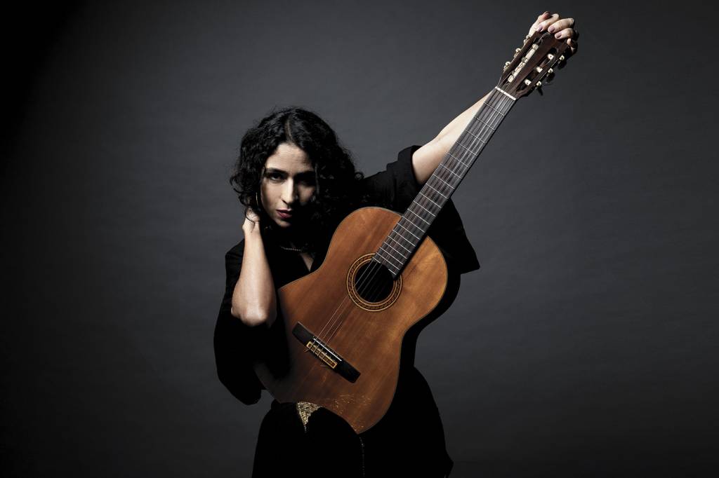 Marisa Monte com vestido preto segura violão e aponta o braço do instrumento para seu lado direito. Ela mira a câmera com olhar desafiador.