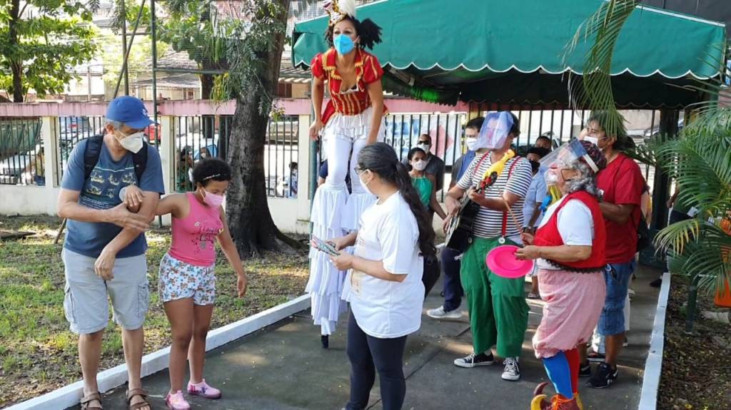 Foto mostra crianças em posto de saúde entretidas por uma artista com perna de pau