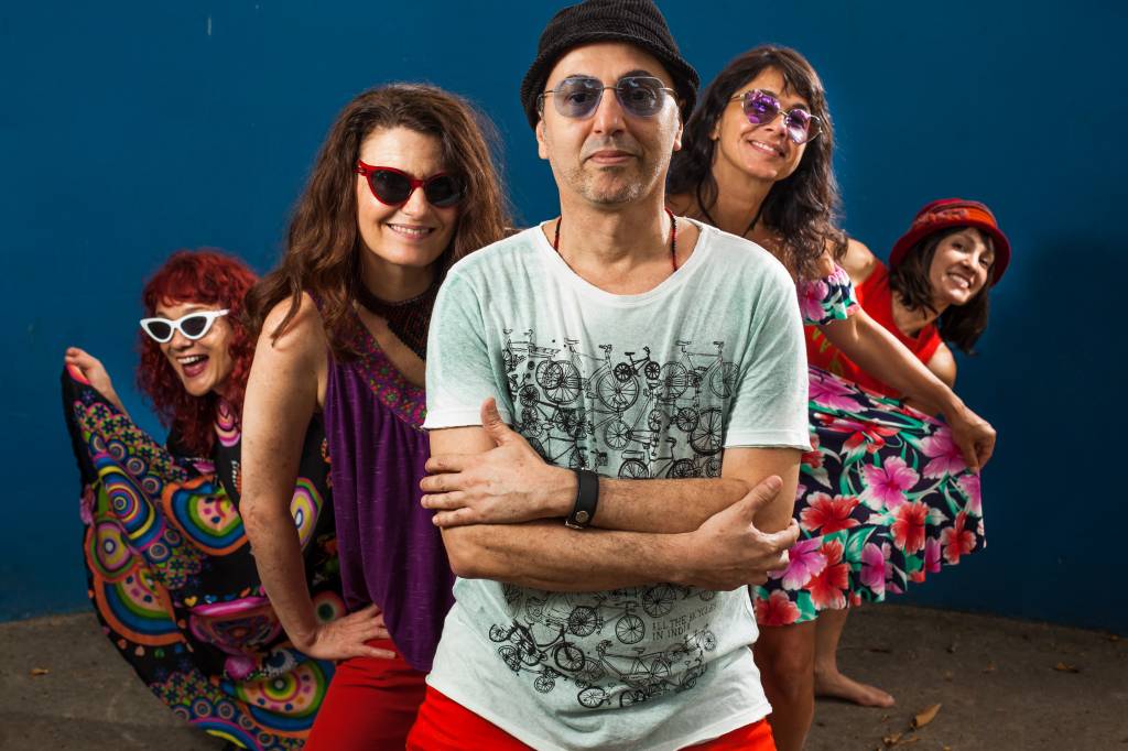 O cantor Zeca Baleiro está na frente, usando óculos de lentes azuis e chapéu, camiseta branca com estampa preta e calça vermelha. Atrás dele, estão quatro mulheres, integrantes do show Zoró Zureta, duas à esquerda e duas à direita.