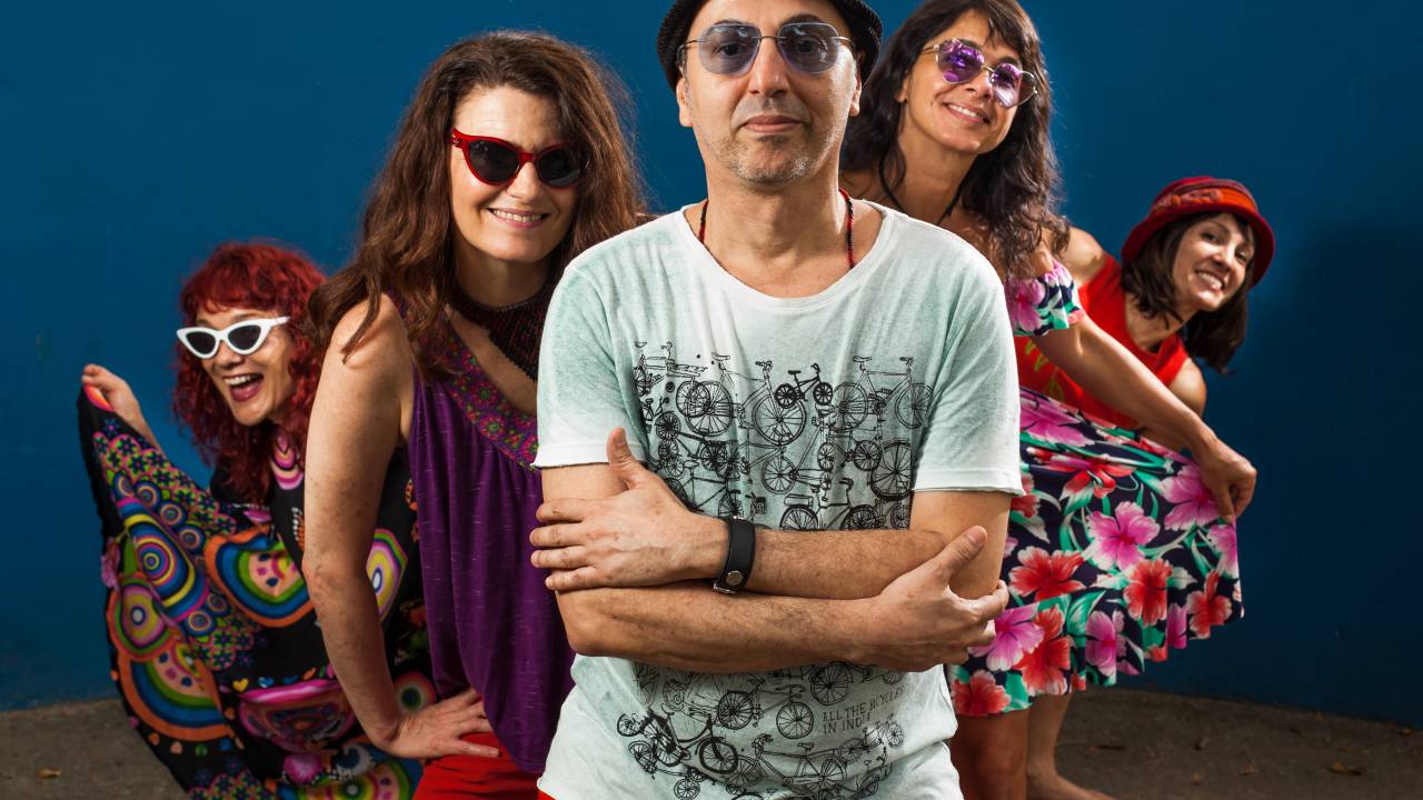 O cantor Zeca Baleiro está na frente, usando óculos de lentes azuis e chapéu, camiseta branca com estampa preta e calça vermelha. Atrás dele, estão quatro mulheres, integrantes do show Zoró Zureta, duas à esquerda e duas à direita.