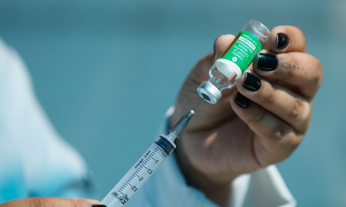 Mão de mulher com unhas pintadas de marrom escuro segurando um frasco de vacina com a mão esquerda e tirando o conteúdo e enchendo com uma seringa com o imunizante na direita