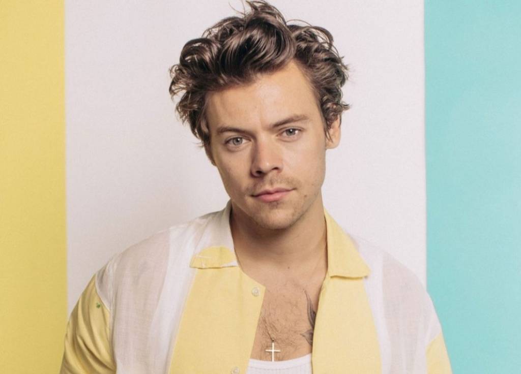 O cantor Harry Styles com penteado meio descabelado. Ele usa camiseta branca e, por cima, camisa com faixas amarelo-claras e brancas na vertical. Ao fundo, uma parede com faixas amarela, branca e azul claro