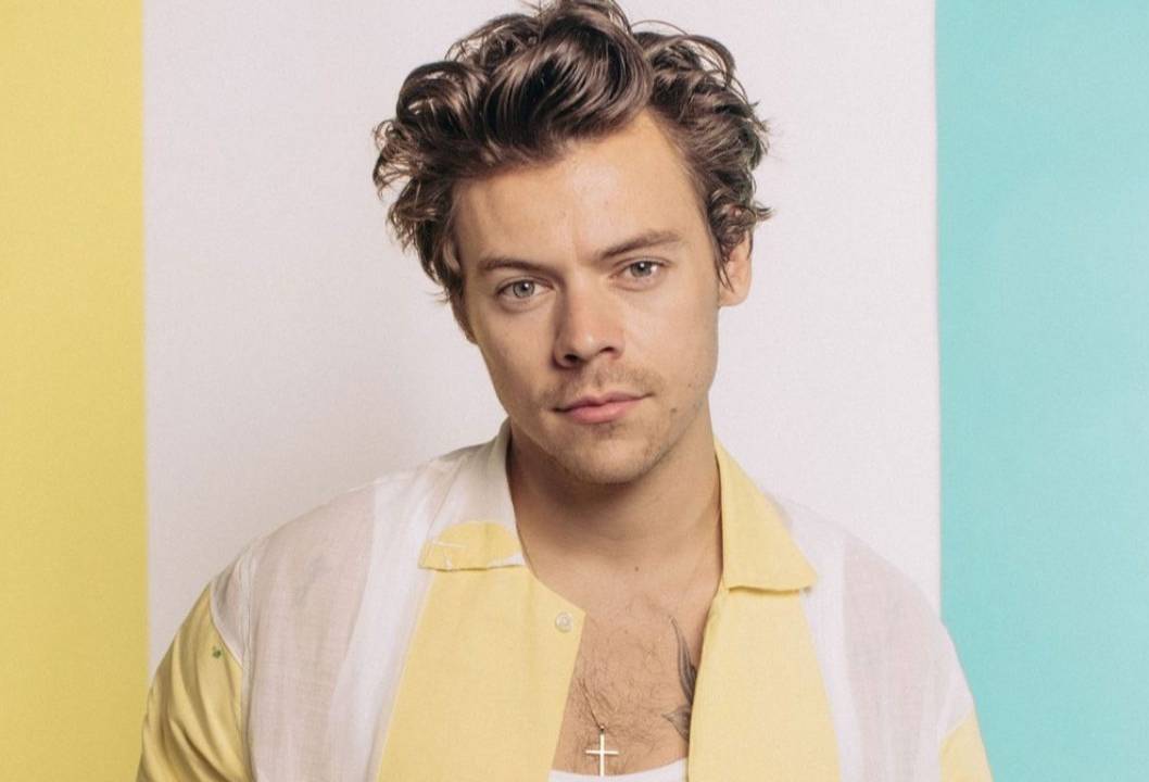 O cantor Harry Styles com penteado meio descabelado. Ele usa camiseta branca e, por cima, camisa com faixas amarelo-claras e brancas na vertical. Ao fundo, uma parede com faixas amarela, branca e azul claro