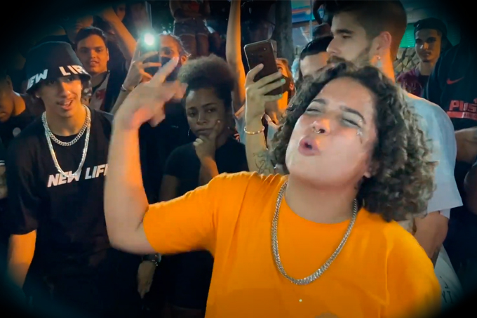 Foto mostra jovem de camisa laranja e cabelo enrolado curto fazendo rimas de rap