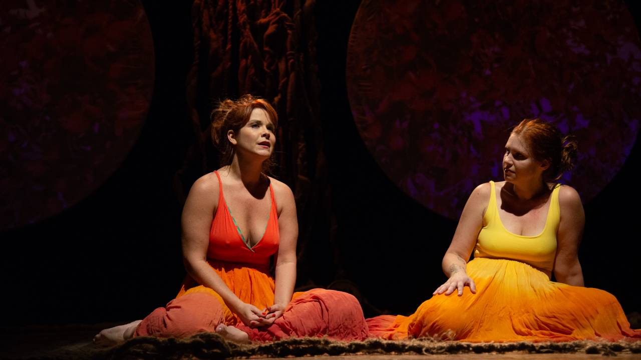 Samara Felippo está sentada e usa vestido de alça com decote laranja. Carolinie Figueiredo está à direita dela e olha pra ela, usando vestido laranja claro.