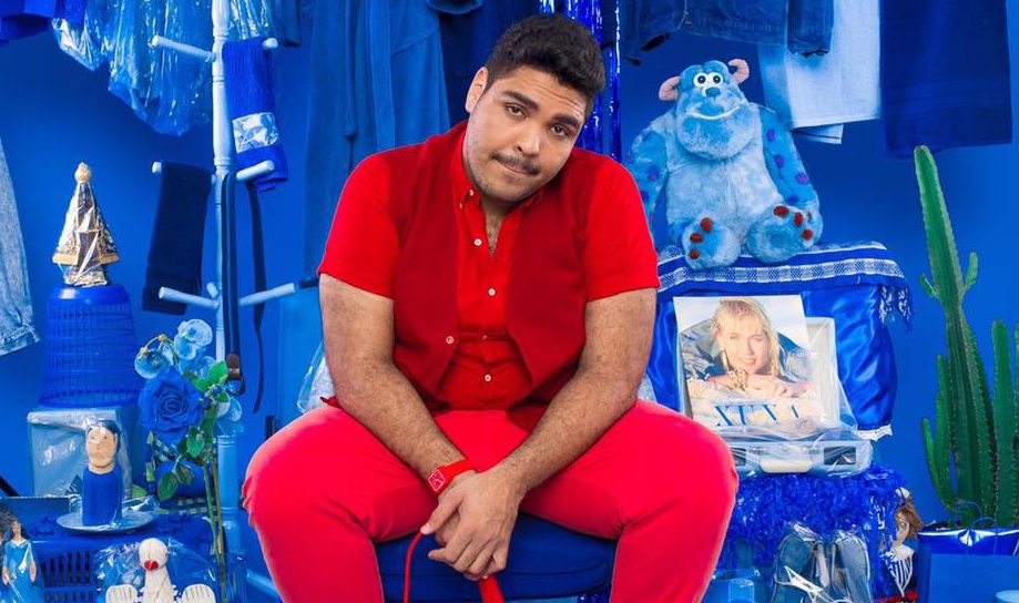 O humorista Paulo Vieira está sentado e usa camisa, calça e colete vermelhos Atrás dele, um fundo azul cheio de objetos azuis.