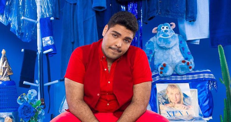 O humorista Paulo Vieira está sentado e usa camisa, calça e colete vermelhos Atrás dele, um fundo azul cheio de objetos azuis.