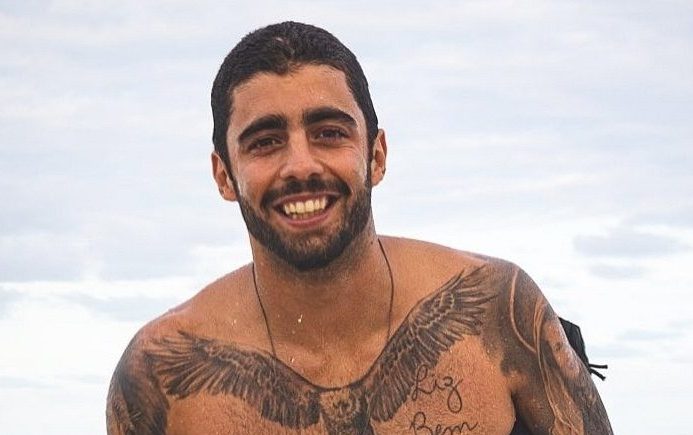 surfista Pedro Scooby saindo do mar, molhado. Ao fundo, o céu azul. Ele tem tatuagens no peito e nos braços. Está de barba e está sorrido.