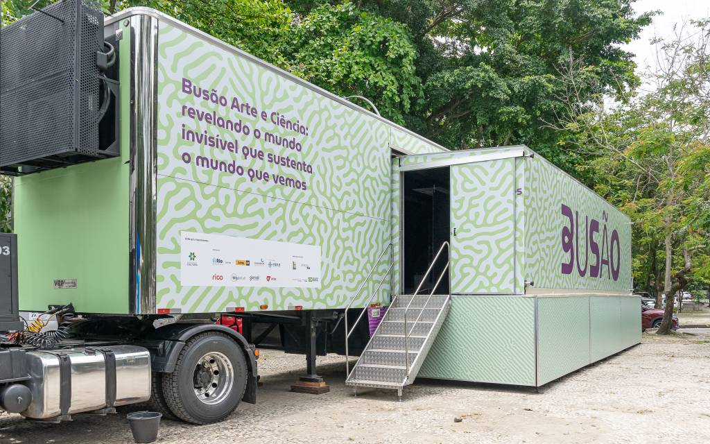 Um caminhão de cor verde-claro estacionado. Na lateral, se lê em roxo: Busão da Arte e Ciência: revelando o mundo invisível que sustenta o mundo em vivemos