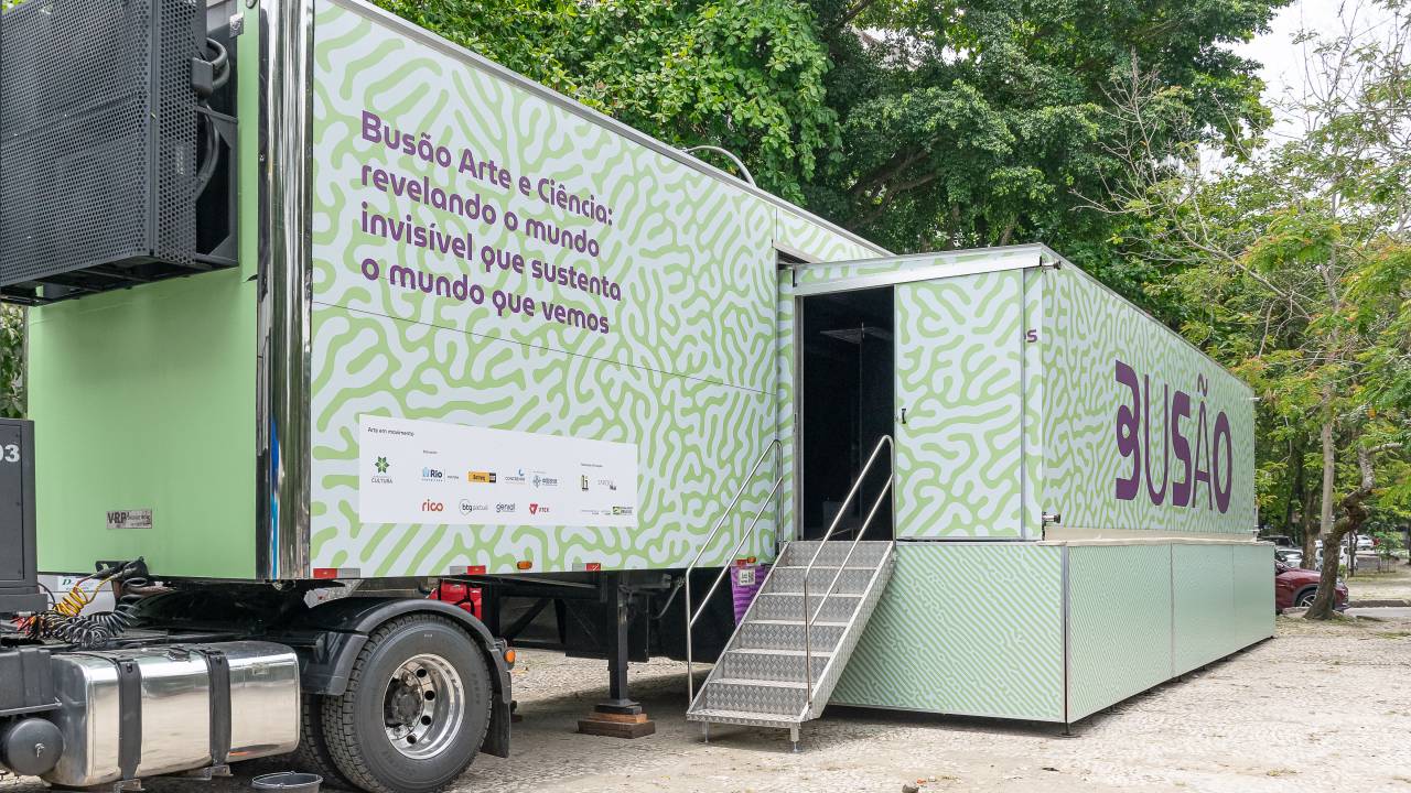 Um caminhão de cor verde-claro estacionado. Na lateral, se lê em roxo: Busão da Arte e Ciência: revelando o mundo invisível que sustenta o mundo em vivemos