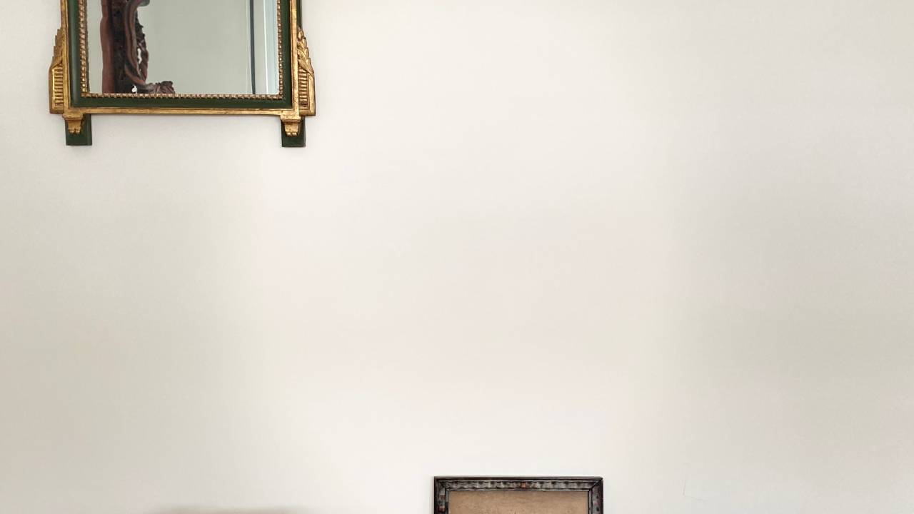 A imagem mostra uma parede com quadros apoiados no chão, um banquinho e um espelho pendurado na parede